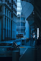 A modern urban scene featuring tall buildings with a lot of glass surfaces. Several cars are parked along the street, and a few people are walking on the sidewalk. The architecture is sleek and contemporary, with an emphasis on straight lines and metallic elements. The setting appears to be a city street with minimal traffic and a few street signs visible.