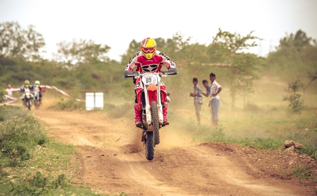 A motorcyclist wearing a vibrant red and yellow helmet and matching suit performs a jump on a dirt trail, with dust kicking up under the bike's spinning wheels. Additional racers follow behind on the rugged track, flanked by greenery and spectators. The scene is set outdoors, characterized by a natural, earthy landscape.