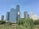 A cluster of modern skyscrapers rises against a clear blue sky, bordered by lush greenery in the foreground. The buildings feature reflective glass windows that catch the sunlight. A construction crane is visible among the high-rises, suggesting ongoing development.