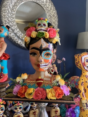 A colorful decorative bust inspired by Dia de los Muertos, featuring a highly detailed face with traditional sugar skull makeup. The bust is adorned with a vibrant floral crown, and various bright elements are incorporated into the design. In the background, other similarly styled sculptures can be seen, adding to the festive and artistic atmosphere.