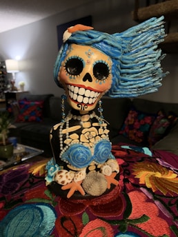 A colorful sculpture of a skeleton figure with vibrant blue hair, large black eye sockets, and a broad smile showing teeth. The skeleton is adorned with elaborate jewelry and seashell decorations, including a top resembling blue roses and starfish on the torso. The background includes a cozy interior setting with a sofa and lamp.