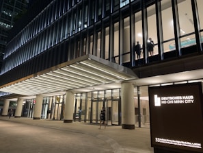 A modern office building with large glass windows, illuminated at night. Two individuals are visible through a window on an upper floor. The entrance is brightly lit, and a signboard displays 'Deutsches Haus Ho Chi Minh City.'