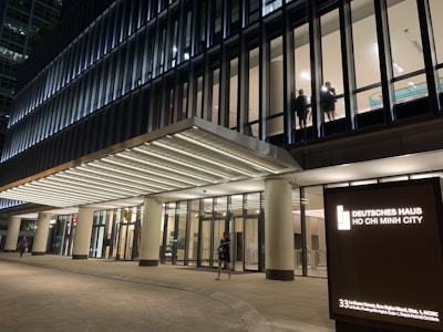 A modern office building with large glass windows, illuminated at night. Two individuals are visible through a window on an upper floor. The entrance is brightly lit, and a signboard displays 'Deutsches Haus Ho Chi Minh City.'