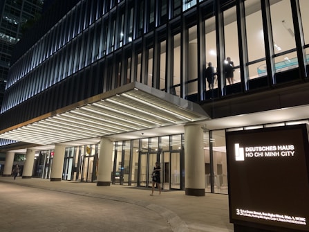 A modern office building with large glass windows, illuminated at night. Two individuals are visible through a window on an upper floor. The entrance is brightly lit, and a signboard displays 'Deutsches Haus Ho Chi Minh City.'