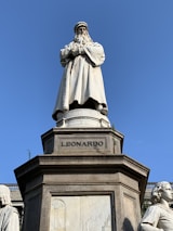 A marble statue of a historical figure standing prominently on a pedestal with the inscription 'LEONARDO'. The figure has a long beard and is dressed in a full-length robe, giving an impression of wisdom and authority. Surrounding the main statue are smaller sculptures and detailed bas-reliefs on the pedestal that add to the grandeur of the monument. The sky is clear and blue, providing a contrasting backdrop to the white marble of the statue.