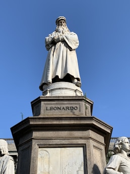 A marble statue of a historical figure standing prominently on a pedestal with the inscription 'LEONARDO'. The figure has a long beard and is dressed in a full-length robe, giving an impression of wisdom and authority. Surrounding the main statue are smaller sculptures and detailed bas-reliefs on the pedestal that add to the grandeur of the monument. The sky is clear and blue, providing a contrasting backdrop to the white marble of the statue.