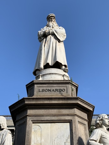 A marble statue of a historical figure standing prominently on a pedestal with the inscription 'LEONARDO'. The figure has a long beard and is dressed in a full-length robe, giving an impression of wisdom and authority. Surrounding the main statue are smaller sculptures and detailed bas-reliefs on the pedestal that add to the grandeur of the monument. The sky is clear and blue, providing a contrasting backdrop to the white marble of the statue.