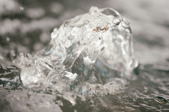 A close-up view of a clear, bubbling water feature. The image captures the texture and movement of the water, highlighting its clarity and fluidity. The background is softly blurred, focusing attention on the central water eruption.