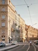 A busy European street scene with classic architecture displaying ornate facades. Several cars and a motorbike travel along the road, and pedestrians walk on the sidewalk. The buildings are multi-storied with tall windows and decorative details. Overhead, tram cables are visible against a clear sky.