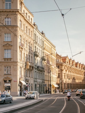 A busy European street scene with classic architecture displaying ornate facades. Several cars and a motorbike travel along the road, and pedestrians walk on the sidewalk. The buildings are multi-storied with tall windows and decorative details. Overhead, tram cables are visible against a clear sky.