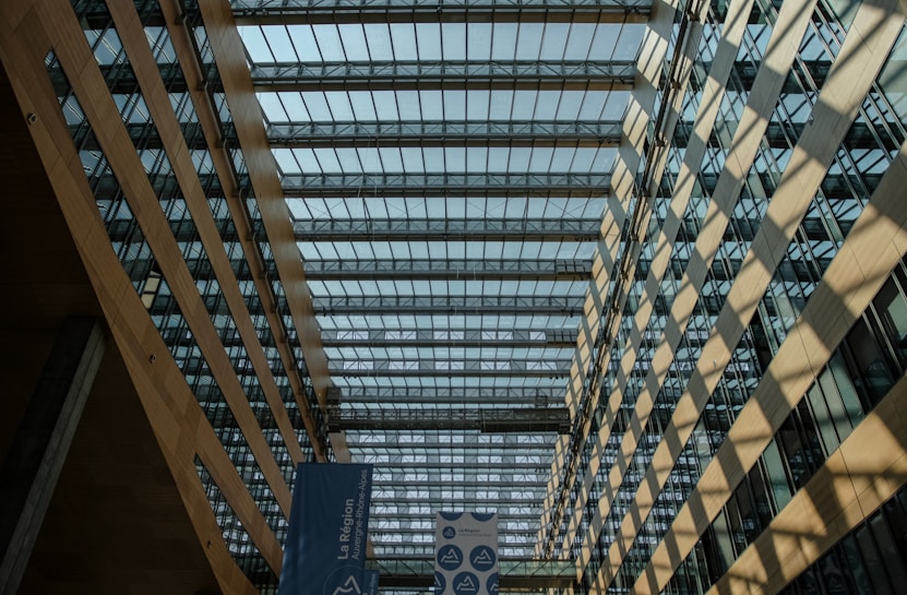 A modern architectural interior featuring a large atrium with extensive glass ceilings and walls. Wooden beams and a geometric pattern of sunlight create shadows on the floor. A blue banner with text is visible in the foreground.