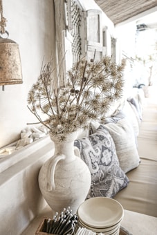 A rustic, neutral-toned interior features a large, textured vase with dried floral arrangements, placed on a mantel or shelf. Decorative pillows with intricate patterns sit on a beige sofa in the background. A wooden wall with weathered, decorative elements and a metal hanging bell are visible, enhancing the cozy, bohemian ambiance.