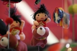 A fabric doll with black hair and a red outfit is riding a plush bear. The background appears blurred, with vibrant colors. The doll has a somewhat neutral expression, and there are other soft objects hanging nearby.