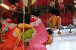 Various plush toys with whimsical designs hang from strings, featuring small figures with black hair and colorful outfits, against a blurred red and white background. Some toys resemble small people riding on larger animal-like figures.