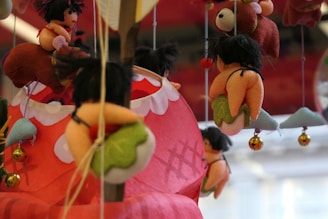 Various plush toys with whimsical designs hang from strings, featuring small figures with black hair and colorful outfits, against a blurred red and white background. Some toys resemble small people riding on larger animal-like figures.