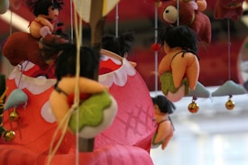 Various plush toys with whimsical designs hang from strings, featuring small figures with black hair and colorful outfits, against a blurred red and white background. Some toys resemble small people riding on larger animal-like figures.