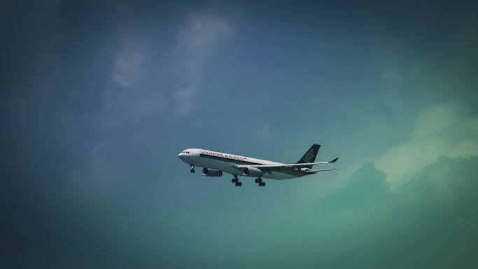 A commercial airplane is flying in the sky, featuring the logo and branding of an airline company on its fuselage. The aircraft is captured against a backdrop of a gradient sky transitioning from a dark to light blue hue with clouds visible.