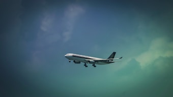 A commercial airplane is flying in the sky, featuring the logo and branding of an airline company on its fuselage. The aircraft is captured against a backdrop of a gradient sky transitioning from a dark to light blue hue with clouds visible.