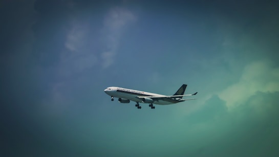 A commercial airplane is flying in the sky, featuring the logo and branding of an airline company on its fuselage. The aircraft is captured against a backdrop of a gradient sky transitioning from a dark to light blue hue with clouds visible.