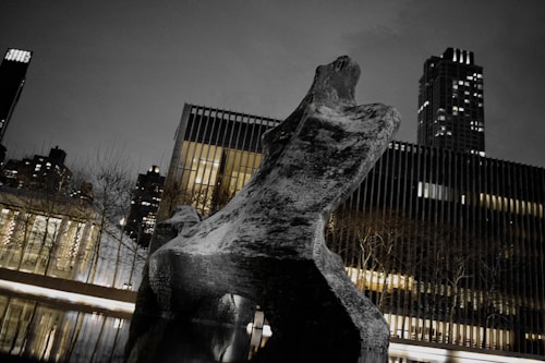 A large abstract sculpture stands prominently in front of modern buildings. The scene is captured at night, with city lights illuminating the surroundings. The sky is dark, enhancing the dramatic presence of the artwork. Reflections from a nearby body of water add to the composition, creating a serene and contemplative atmosphere.