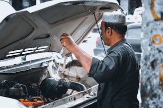 A man wearing a traditional cap is working under the hood of a vehicle. He appears to be inspecting or fixing the engine, with various engine components visible in the foreground. The background is slightly blurred, suggesting a street or outdoor setting.