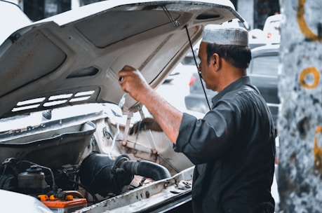 A man wearing a traditional cap is working under the hood of a vehicle. He appears to be inspecting or fixing the engine, with various engine components visible in the foreground. The background is slightly blurred, suggesting a street or outdoor setting.
