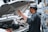 A man wearing a traditional cap is working under the hood of a vehicle. He appears to be inspecting or fixing the engine, with various engine components visible in the foreground. The background is slightly blurred, suggesting a street or outdoor setting.
