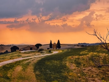 A picturesque countryside scene with rolling hills and a dirt road leading to a rustic farmhouse surrounded by trees. The sky features dramatic orange and purple hues from a sunset or sunrise, adding warmth and depth to the image.