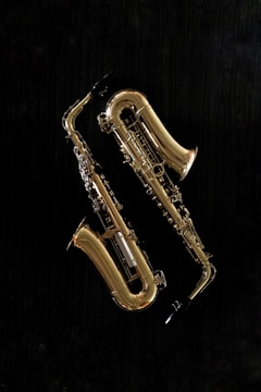 Two brass saxophones are displayed against a solid black background. The instruments are gleaming, revealing intricate details and the curves of the saxophone body. The lighting highlights the shiny, reflective surface of the brass.