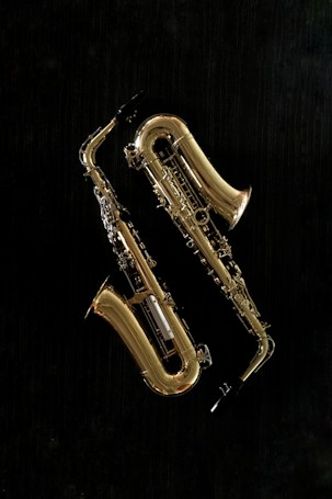 Two brass saxophones are displayed against a solid black background. The instruments are gleaming, revealing intricate details and the curves of the saxophone body. The lighting highlights the shiny, reflective surface of the brass.