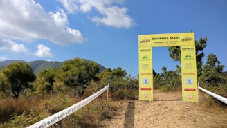 A yellow archway marks the start of a downhill biking trail in a mountainous area. The archway displays information about the Kathmandu MTB Fest 2016. The scene is surrounded by green shrubs and trees under a partly cloudy sky.
