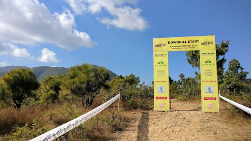 A yellow archway marks the start of a downhill biking trail in a mountainous area. The archway displays information about the Kathmandu MTB Fest 2016. The scene is surrounded by green shrubs and trees under a partly cloudy sky.