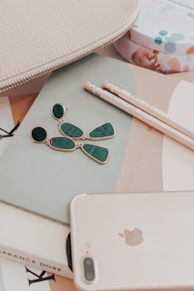 A flat lay featuring a smartphone, two pencils, a pair of dangling green earrings, several books, and a textured beige bag. The composition is aesthetically arranged, highlighting daily essentials and accessories.