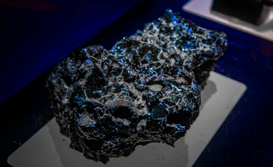 A rough, jagged black rock with glistening blue reflections sits on a display platform. The surface is uneven with multiple indentations and shiny patches that catch the light.