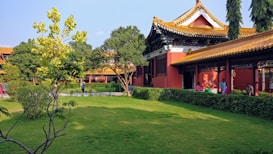 A serene garden scene featuring traditional Asian architecture with red and gold tones. The building has ornate roof designs and is surrounded by lush green grass and trees. People are sitting and walking around the garden, enjoying the peaceful environment.