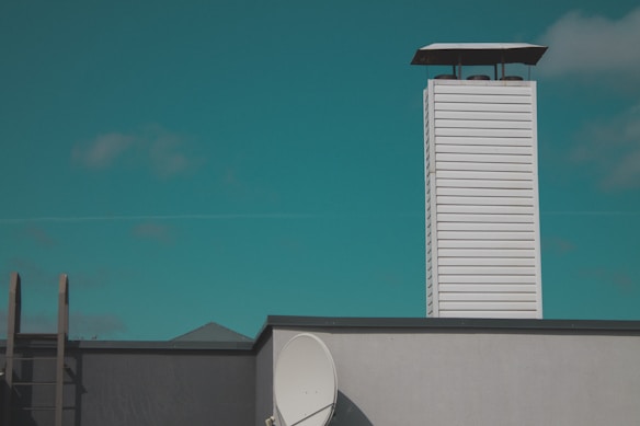 A rooftop scene featuring a tall white vent or chimney with horizontal slats, set against a clear teal sky. A satellite dish is attached to the side of the building, and there is a ladder leaning against the roof.