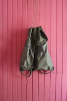 A green backpack hangs against a vertically paneled pink wooden wall. The backpack has leather straps and an overall rugged appearance, suggesting it is meant for outdoor use.
