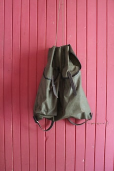 A green backpack hangs against a vertically paneled pink wooden wall. The backpack has leather straps and an overall rugged appearance, suggesting it is meant for outdoor use.
