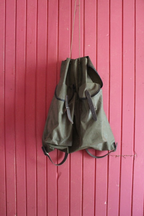 A green backpack hangs against a vertically paneled pink wooden wall. The backpack has leather straps and an overall rugged appearance, suggesting it is meant for outdoor use.