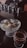 A glass dish filled with several scoops of vanilla ice cream is accompanied by a delicate glass containing a shot of espresso. The background features a reflective surface with various coffee-making implements, including metallic containers.