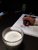A hand is drawing a sketch of a person on a white sheet of paper using a pen. In the foreground, there is a glass filled with a creamy beverage. The setup suggests a casual and creative environment, potentially a café, as there's also a container of green coffee in the background.