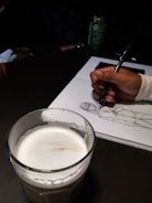 A hand is drawing a sketch of a person on a white sheet of paper using a pen. In the foreground, there is a glass filled with a creamy beverage. The setup suggests a casual and creative environment, potentially a café, as there's also a container of green coffee in the background.