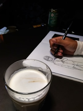A hand is drawing a sketch of a person on a white sheet of paper using a pen. In the foreground, there is a glass filled with a creamy beverage. The setup suggests a casual and creative environment, potentially a café, as there's also a container of green coffee in the background.
