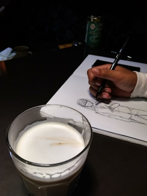 A hand is drawing a sketch of a person on a white sheet of paper using a pen. In the foreground, there is a glass filled with a creamy beverage. The setup suggests a casual and creative environment, potentially a café, as there's also a container of green coffee in the background.
