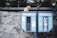A ginger cat sits atop two old, blue public telephone booths that are attached to a stone wall. The booths are labeled 'Türk Telekom' and show signs of wear and graffiti. The surrounding area features greenery and a rugged stone texture.