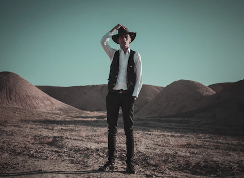 Tony of Tucson standing against a rugged desert backdrop, dressed in classic western attire with his long white beard and stoic expression.