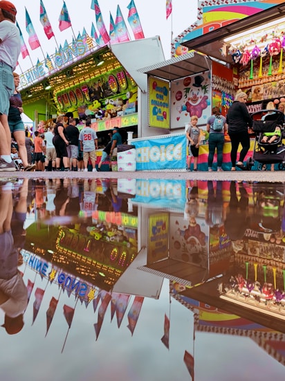 A vibrant carnival scene featuring a variety of game stands and attractions decorated with colorful banners and flags. People can be seen queuing up, interacting, and moving around the stalls that offer various prize games. The ground is wet, creating a reflective puddle that mirrors the lively atmosphere above.