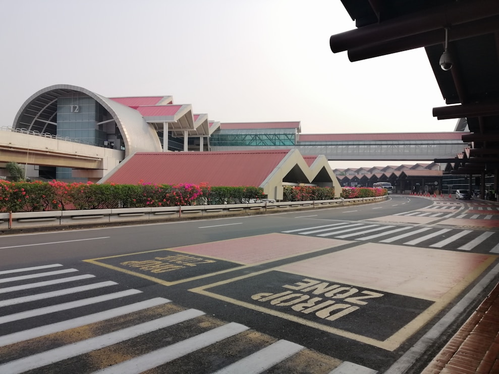 A modern airport terminal with distinctive architectural features, including large glass facades and curving roofs. The foreground includes a designated drop-off zone marked on the road, alongside pedestrian crosswalks. The area is landscaped with flowerbeds, adding aesthetic value.