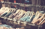 A clothing rack filled with various knitted sweaters hanging on white hangers. The sweaters are arranged closely together and display a range of colors including blue, pink, beige, and green. The rack is metallic, and the background is softly blurred, focusing attention on the clothing items.