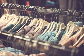 A clothing rack filled with various knitted sweaters hanging on white hangers. The sweaters are arranged closely together and display a range of colors including blue, pink, beige, and green. The rack is metallic, and the background is softly blurred, focusing attention on the clothing items.